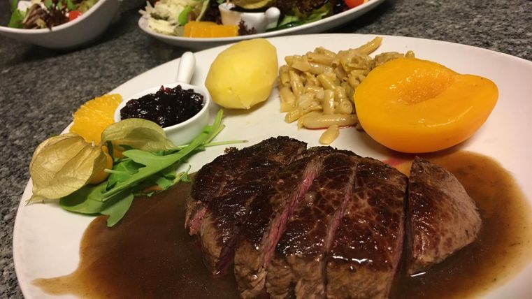 Een bord met sappige steak in donkere saus, geserveerd met aardappelen, perzik en verschillende bijgerechten. Op de achtergrond zijn verse salades te zien.
