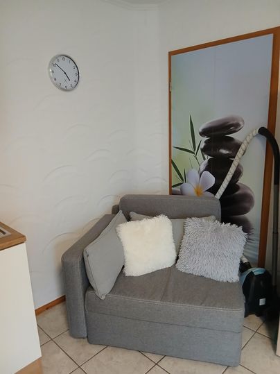 Ein gemütlicher grauer Sessel mit Kissen steht neben einer Wand mit einem entspannenden Motiv von Steinen und Pflanzen. Im Hintergrund hängt eine Uhr an der Wand.