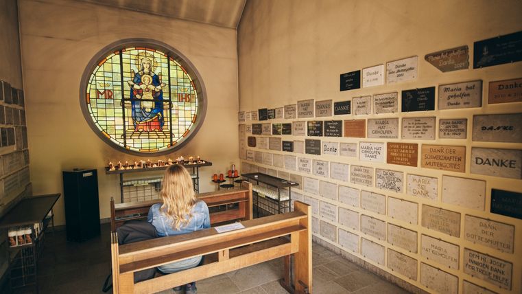 The interior of a small chapel. A person is sitting on a wooden bench. 