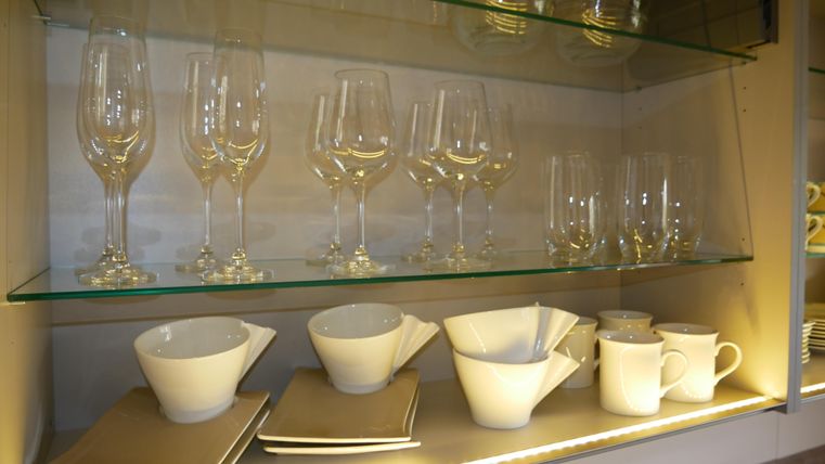 Een reeks glazen en servies in een modern glazen rek. De witte kopjes en kommen zijn netjes gerangschikt en goed zichtbaar.