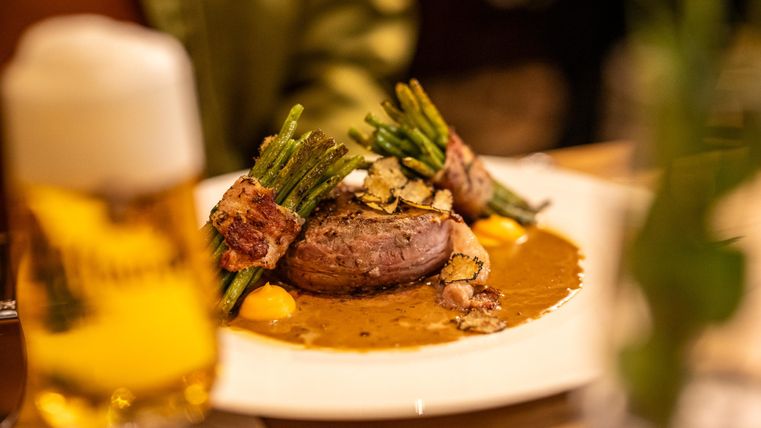 Ein Teller mit saftigem Fleisch, umwickelt mit Speck und Spargel, auf einer cremigen Sauce. Im Hintergrund steht ein Glas Bier.