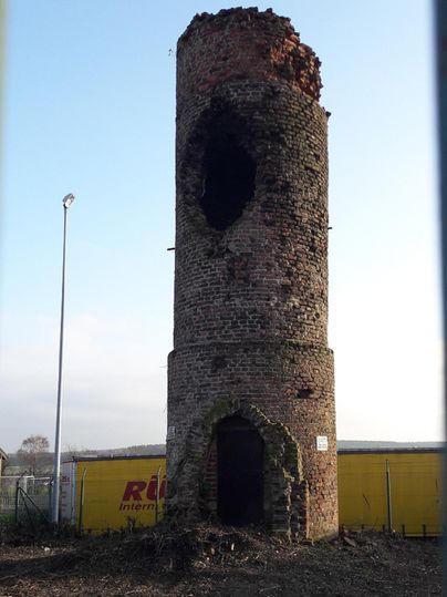 An old, damaged brick tower stands in an open landscape. The upper part is severely dilapidated and the sky in the background is clear.
