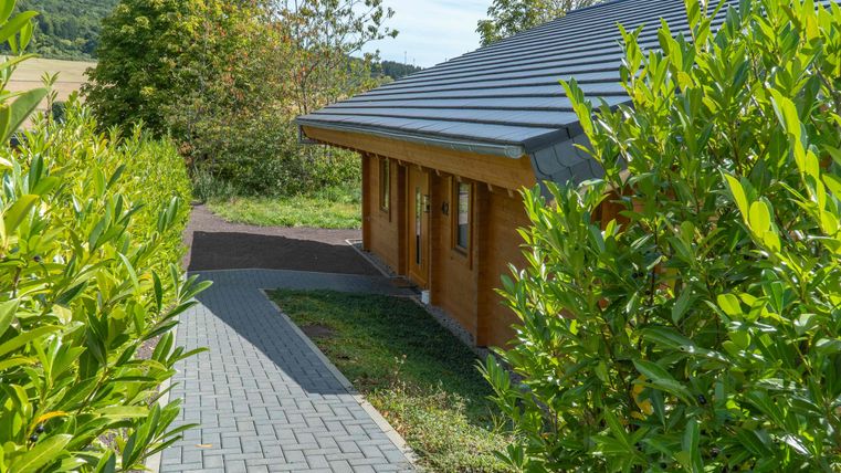 Ein Holzhaus mit einem modernen Dach, umgeben von grünen Sträuchern. Ein gepflasterter Weg führt zu dem Haus in einer ländlichen Umgebung.