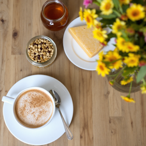 Een kopje koffie op een witte schotel, vergezeld van honing, kaas en een kom met graan. Daarnaast staat een vaas met gele bloemen.