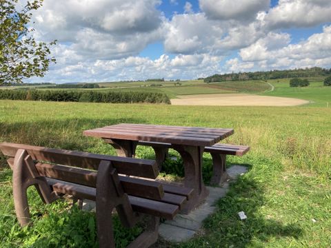 Wooden table and benches on a meadow with a wide view over fields and hills under a cloudy sky.