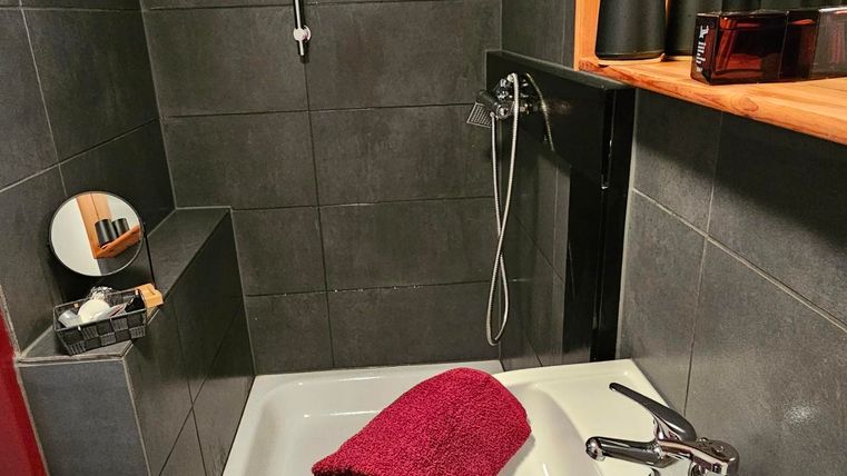 A modern bathroom with a shower and a bathtub. There are some towels and a wooden shelf with care products.
