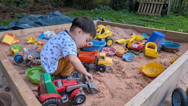 Ein kleines Kind spielt in einer Sandkiste voller bunter Spielzeuge. Es gräbt mit einem Schaufel im Sand und umgeben von Traktoren und anderen Baufahrzeugen.