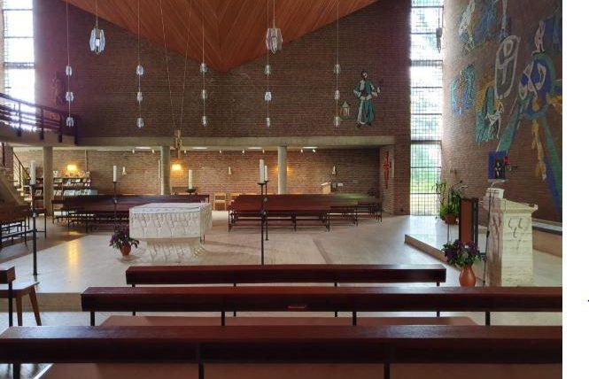 Eine moderne Kirche mit einem hellen Innenraum und einer auffälligen Holzdecke. Im Vordergrund stehen Bänke, und im Hintergrund ist ein Altar sowie bunte Wandmalereien zu sehen.
