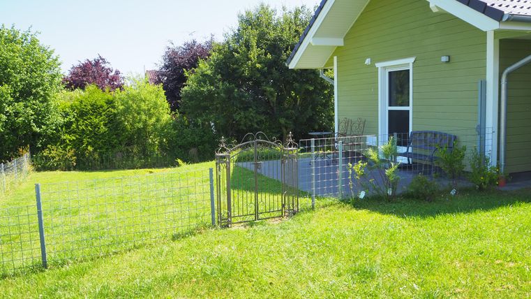 Ein schönes, grünes Grundstück mit einem kleinen Gartenzaun. Im Hintergrund steht ein hellgrünes Haus mit einem gemütlichen Sitzbereich.