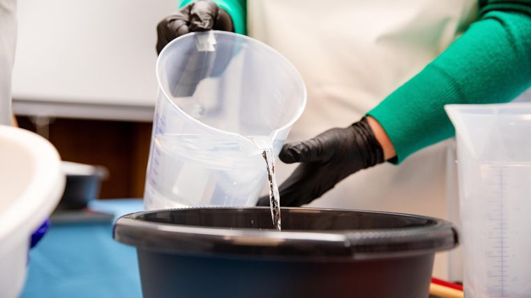 A person is pouring water from a measuring cup into a black bucket. The hands are covered with black gloves.