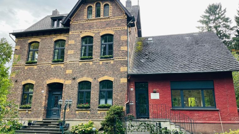 Ein historisches Gebäude mit roten und braunen Ziegeln. Der Eingangsbereich ist von einem gepflegten Garten umgeben.