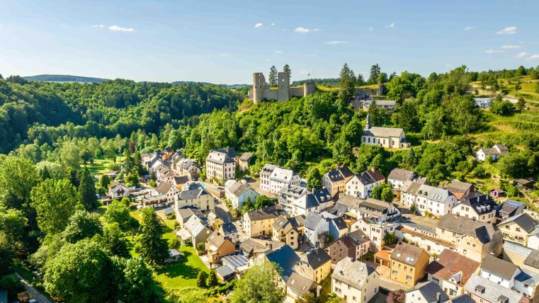 Un paysage pittoresque avec un petit village et un château en arrière-plan. Un vert luxuriant et un ciel ensoleillé créent une atmosphère idyllique.