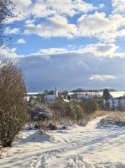 Un paysage enneigé avec un petit village et une église en arrière-plan. Le ciel est partiellement nuageux et diffuse une atmosphère calme d'hiver.