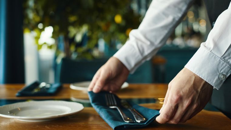 Ein Kellner deckt einen Tisch mit Besteck und einer Serviette. Der Tisch ist elegant und gut vorbereitet für Gäste.