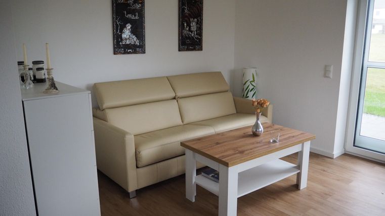 Ein moderner Wohnraum mit einem beigen Sofa und einem Holzkaffetisch. An den Wänden hängen zwei Bilder, und große Fenster sorgen für viel Tageslicht.