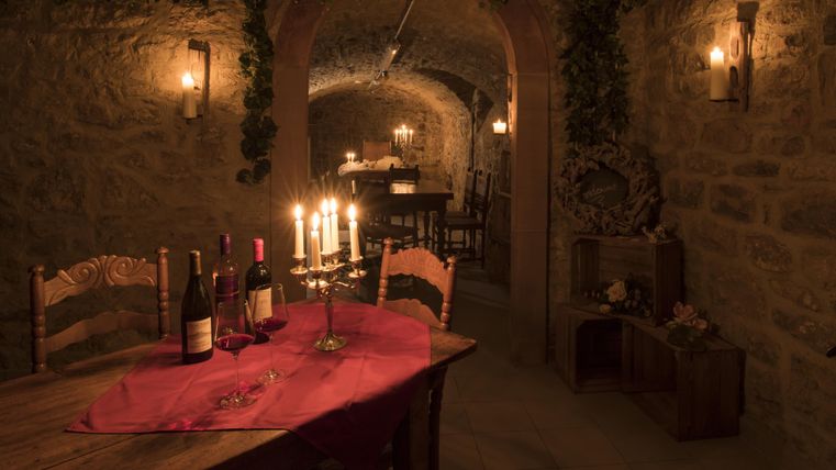Ein gemütlicher Weinkeller mit Steinmauern und Kerzenlicht. Auf einem Tisch stehen Weinflaschen und eine rote Tischdecke.