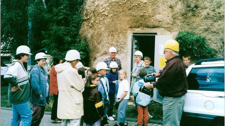 Eine Gruppe von Kindern und Erwachsenen steht vor einem Eingang zu einer Höhle. Alle tragen Helme und hören einem Mann zu, der Informationen gibt.