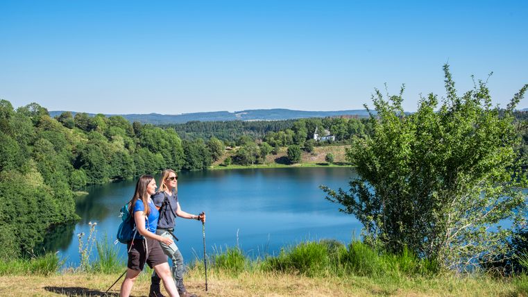 Two hikers are walking on a hill alongside a small lake. In the background is a hilly, wooded landscape from which a small chapel stands out.
