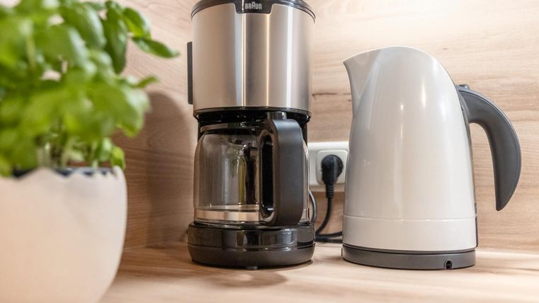 A modern coffee machine stands next to a kettle on a wooden surface. In the background, a pot with fresh herbs can be seen.