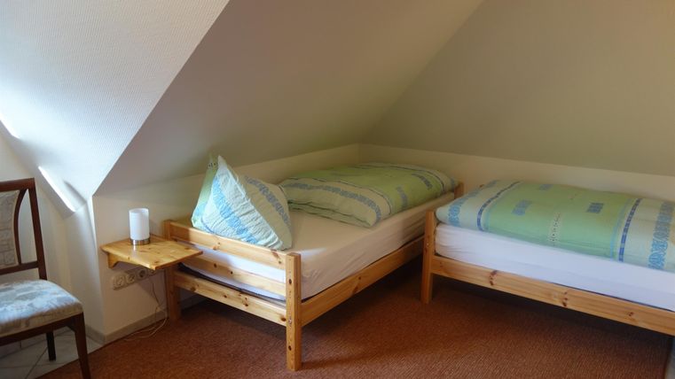 A simple bedroom with two single beds and a chair. The walls are light and the floors are carpeted.