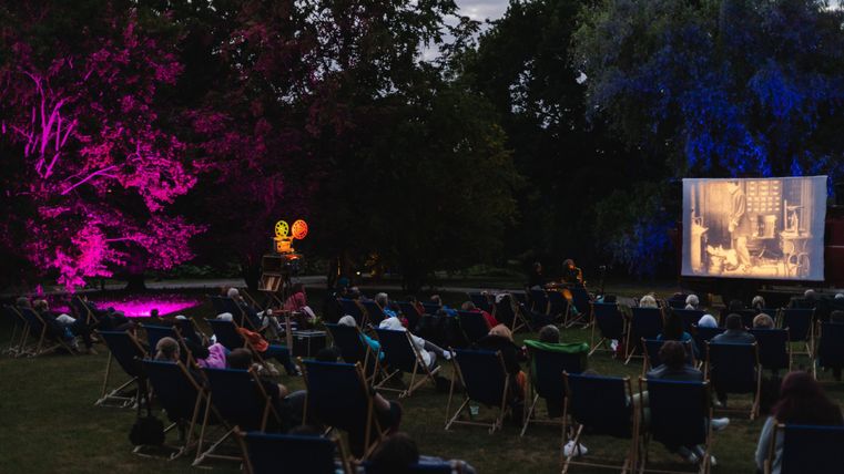Ein Freiluftkino mit vielen Sitzgelegenheiten und einer Leinwand, auf der ein Film gezeigt wird. Bunte Lichter beleuchten die Umgebung und schaffen eine gemütliche Atmosphäre.