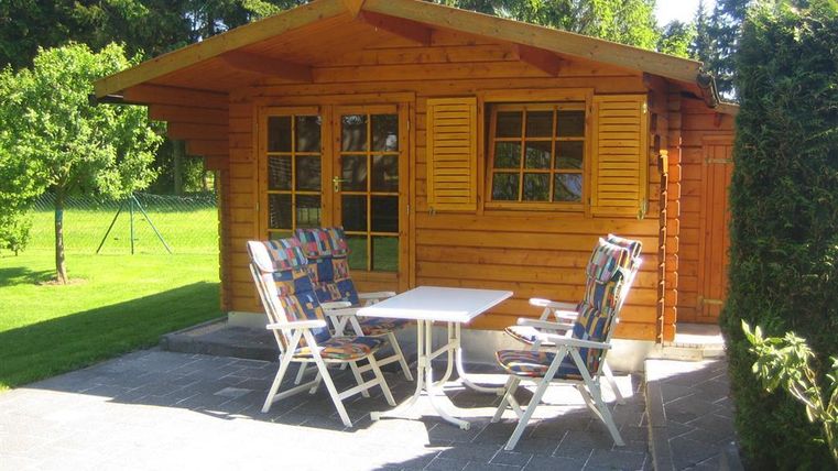 A cozy wooden house with a small terrace and two garden chairs. The garden is green and brightly lit by the sun.