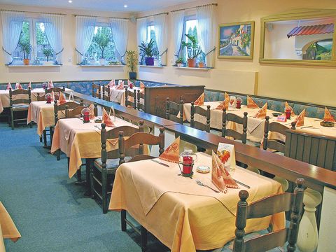 Meerdere tafels zijn versierd met een licht-oranje tafellaken, oranje servetten en rode kandelaren, aan de muren hangen schilderijen en op de vensterbanken staan planten