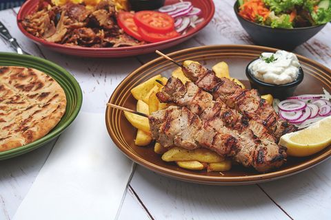 On a wooden table are four dishes: gyros with fries, meat, bread, and a mixed salad