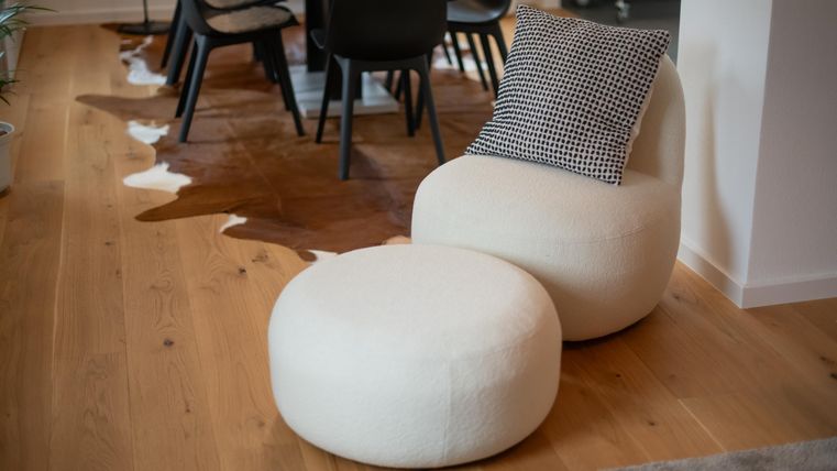 Ein moderner Raum mit einem bequemen Stuhl und einem runden Pouf in hellen Farben. Der Boden ist aus Holz und es gibt einen Esstisch im Hintergrund.