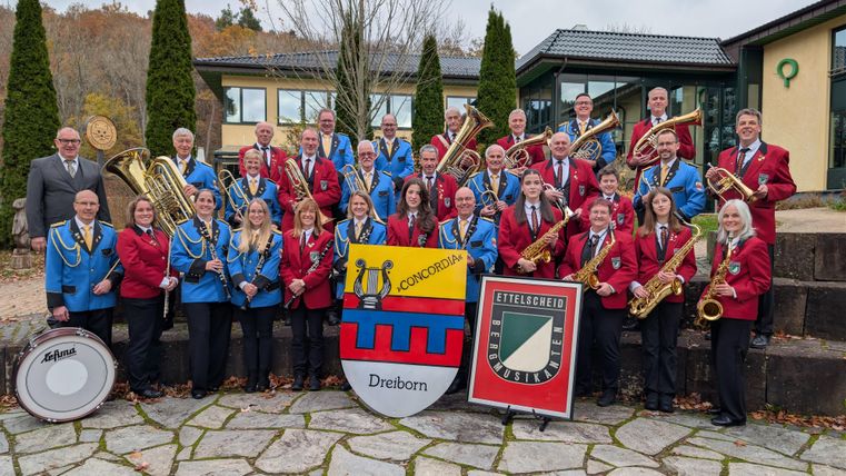 Ein Platz mit einem Orchester, das unter einem überdachten Bereich auftritt. Im Vordergrund steht ein Banner mit dem Logo von Ettelscheid.
