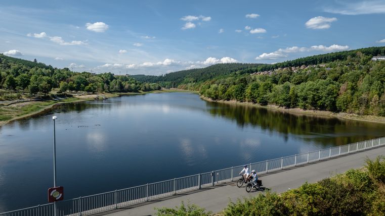 Two cyclists ride on a bridge over Lake Kronenburg in slightly cloudy weather. In the background, Lake Kronenburg is surrounded by a wooded area, from which a village juts out on the right-hand side.