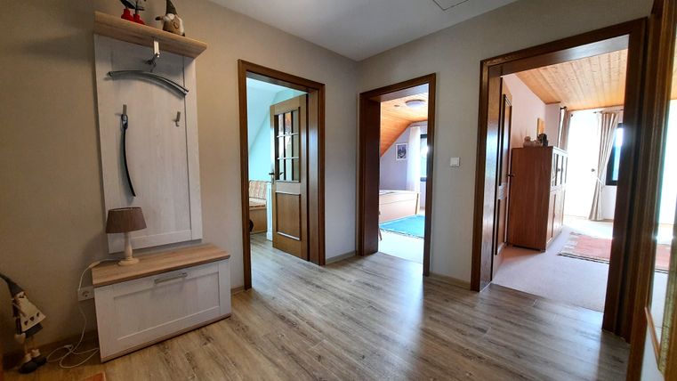 Un couloir lumineux avec un sol en bois et plusieurs portes menant à d'autres pièces. Les murs sont peints dans des couleurs neutres et il y a un petit placard en bois.