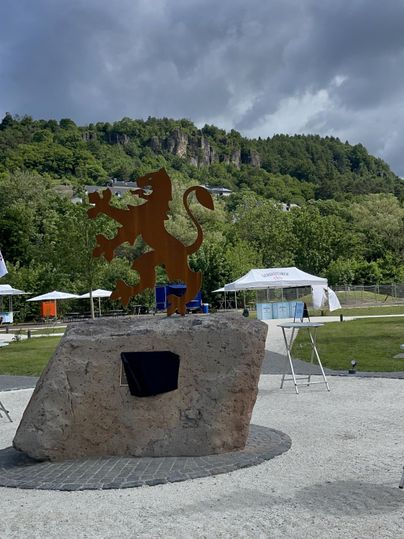 Een roestige metalen leeuwstandbeeld staat op een grote kei voor een beboste heuvelrug. Op de voorgrond en achtergrond zijn tenten en tafels te zien, wat op een evenement wijst.