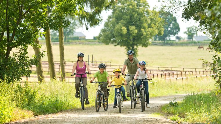 Une famille circule à vélo sur un chemin rural entouré d'arbres et de prairies.
