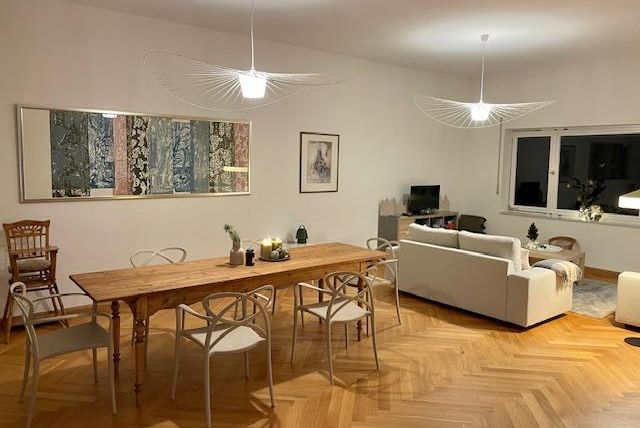 Ein modernes Wohnzimmer mit einem langen Holztisch und hellen Stühlen. Im Hintergrund sind ein gemütliches Sofa und große Fenster zu sehen.
