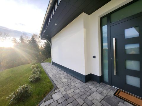 Een moderne entree van een huis met een grote glazen front. Het pad is verhard en omgeven door een groene gazon, terwijl de zon op de achtergrond opkomt.