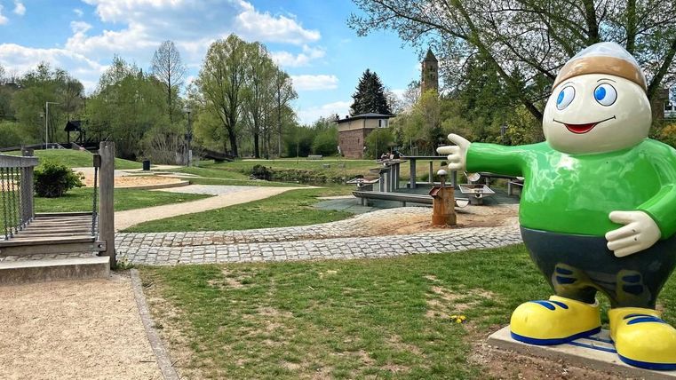 A colorful figure stands in a park with green meadows and paths. In the background, there are trees and a small stream.