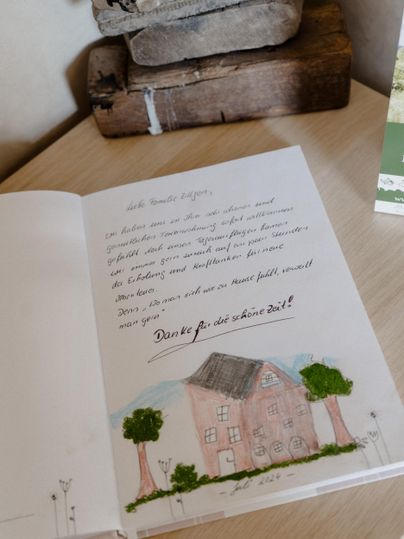 Ein offenes Buch mit einer handgeschriebenen Nachricht und einer Zeichnung eines Hauses umgeben von Bäumen. Es steht auf einem Tisch neben einem Holzblock und anderen dekorativen Objekten.