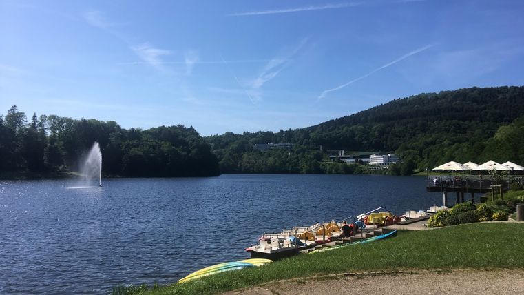 Lac de barrage de Bitburg avec pédalos et fontaines, entouré d'arbres et de collines.