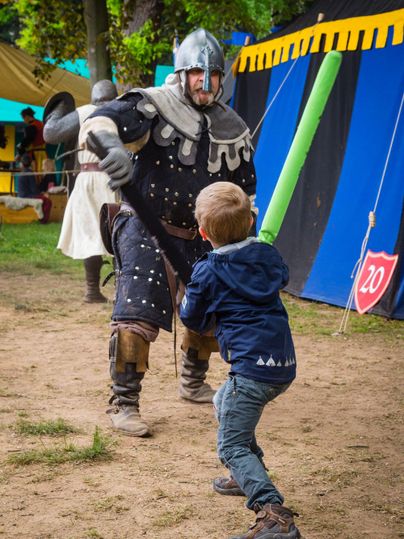 Ein Ritter in Rüstung kämpft mit einem Jungen, der ein Lichtschwert hält. Im Hintergrund sind bunte Zelte zu sehen.