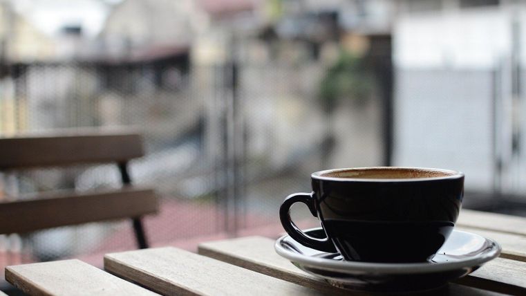 Schwarze Kaffeetasse auf einem Holztisch im Freien, unscharfer Hintergrund mit Gebäuden.