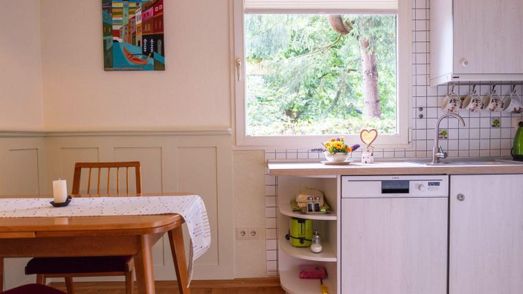 Une cuisine lumineuse avec une grande baie vitrée offrant une vue sur le jardin. Une table à manger avec un chemin de table blanc et une bougie complète la pièce.