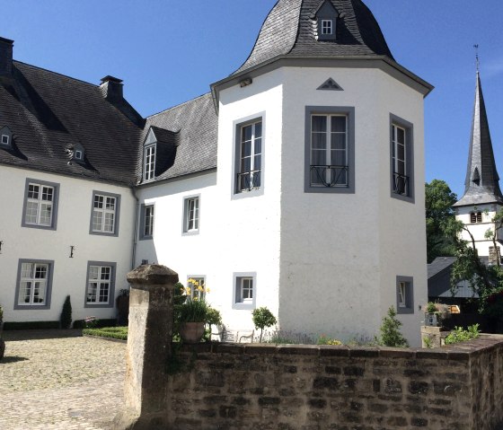 Château blanc avec un toit gris et une tour, entouré d'un jardin et d'un mur de pierre, sous un ciel bleu., © Fellmann / TI Bitburger Land