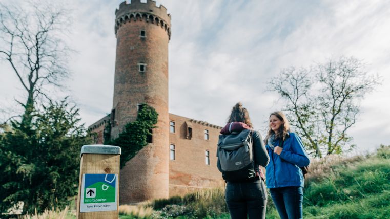 Two people stand in front of a historic tower in Zülpich, with a signpost for the Eifelspur in the foreground.