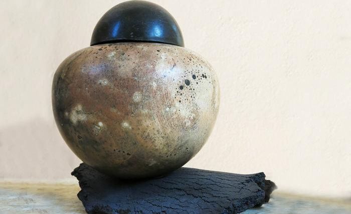 A decorative ceramic vase with a round, smooth surface and a black lid. It stands on a black base and has a softened, antique appearance.
