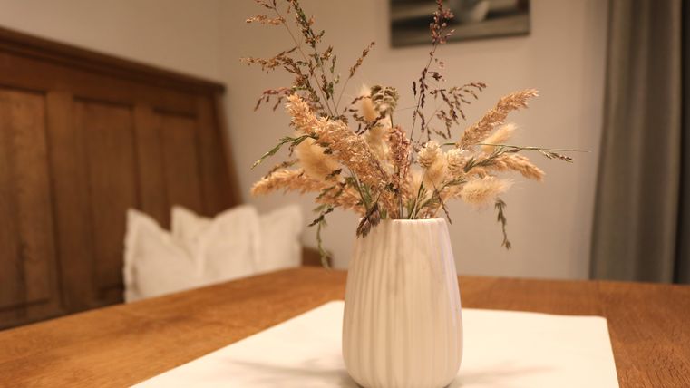 Une élégante vase avec des fleurs séchées se trouve sur une table.
En arrière-plan, des surfaces en bois et une décoration discrète sont visibles.