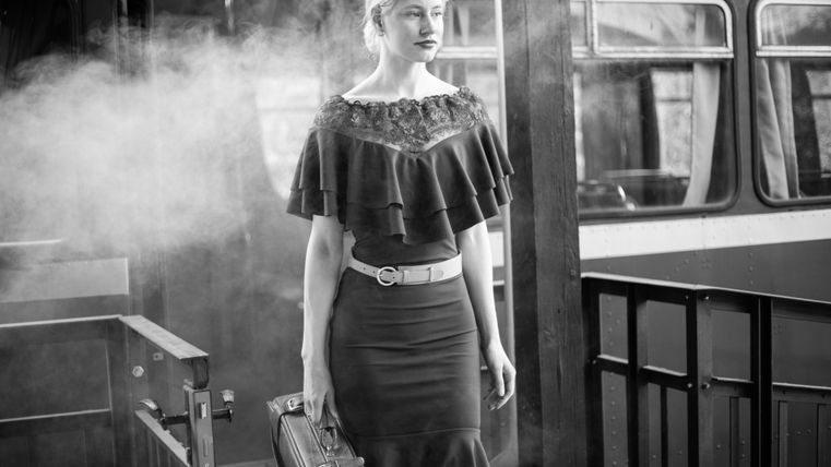 Une femme élégante avec une mallette se tient dans une gare au style rétro. Elle porte une tenue chic et sombre et dégage une impression de confiance.