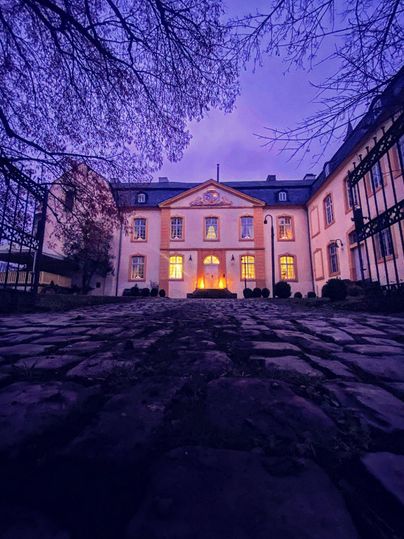 Ein beeindruckendes Gebäude bei Dämmerung mit warmem Licht in den Fenstern. Der Weg aus Kopfsteinpflaster führt zur Eingangstür.