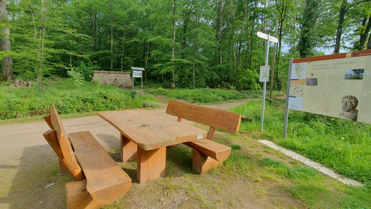 Eine Holzsitzgruppe in der Umgebung von bewaldeter Fläche und einigen Informationstafeln 
