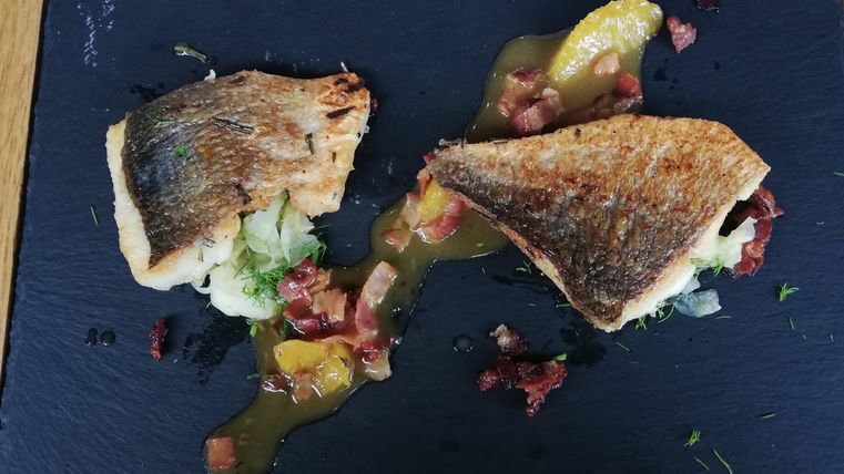 Ein schön angerichtetes Gericht mit gebratenem Fisch auf einem schwarzen Stein. Dazu sind Gemüse und eine besondere Sauce serviert.